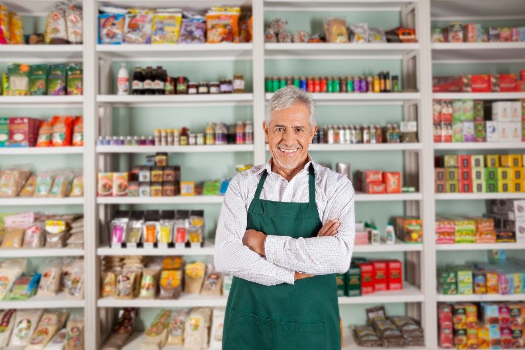 Portrait of happy senior male owner standing arms crossed against shelves